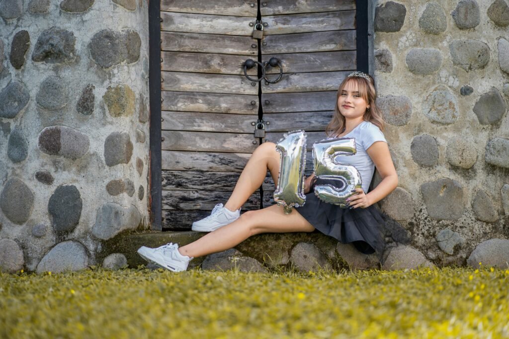 Teen girl in a skirt holding balloons with number 15, sitting by a stone wall.