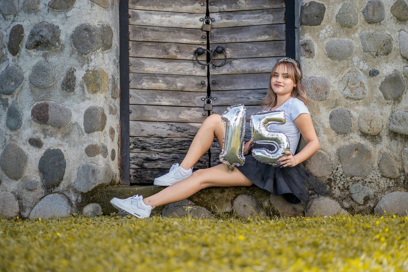 Teen girl in a skirt holding balloons with number 15, sitting by a stone wall.